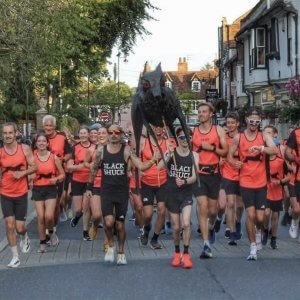 Members of Bungay Black Shuck Running Club running into Bungay with the effigy of the black shuck in 2025. Taken from the from the from two runners are wearing black vests with BLACK SHUCK across the front.